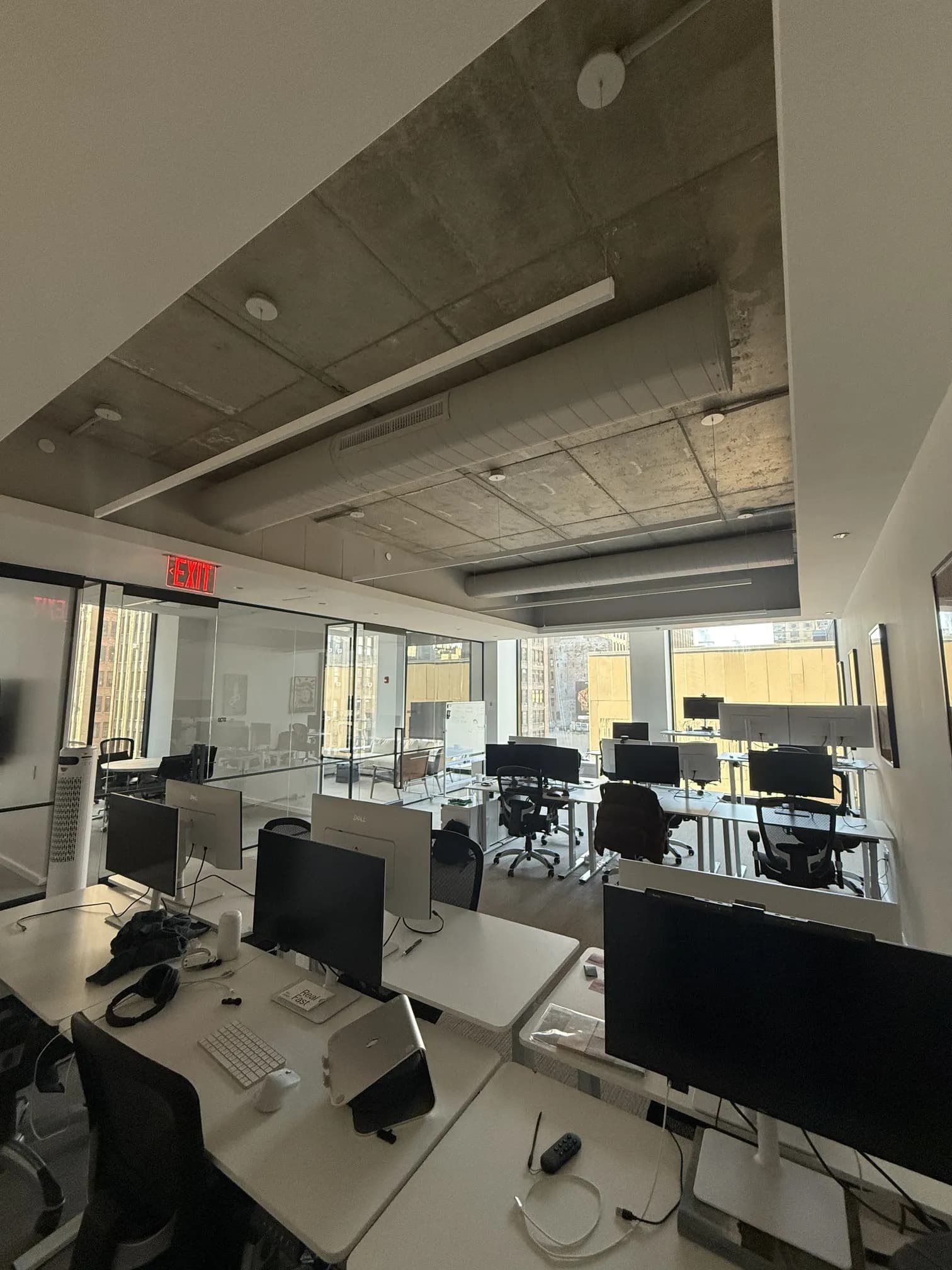 The Melius open-plan office with rows of desks and monitors beneath an exposed concrete ceiling and city-view windows.